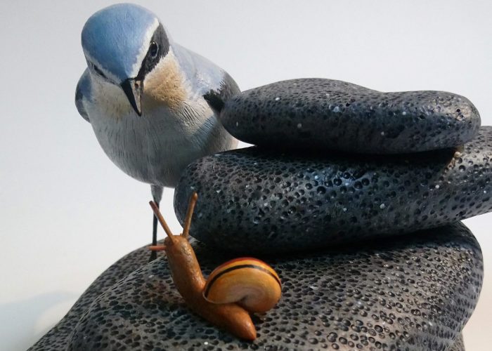Moorland meeting: A painted woodcarving of a wheatear bird looking at a snail on a pile of stones.