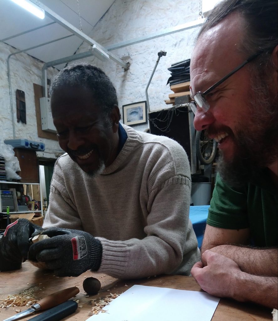 Clarke Peters is carving a small sculpture with a knife and smiling. Alistair Park looks on