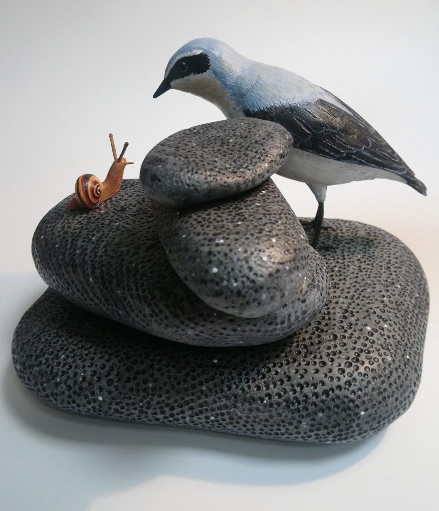 Painted woodcarving of a wheatear bird looking at a banded snail on a pile of stones